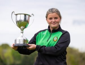 Young woman in a green and black sports jacket holding a large silver trophy outdoors, smiling at the camera.