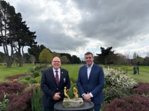 Two men in suits pose beside a bronze statue of two golfers on a stone pedestal in a manicured park garden act as a ceremony backdrop.