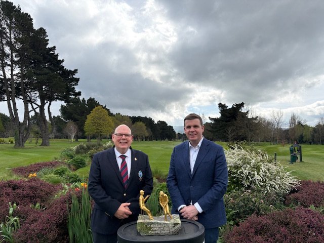 Two men in suits pose beside a bronze statue of two golfers on a stone pedestal in a manicured park garden act as a ceremony backdrop.