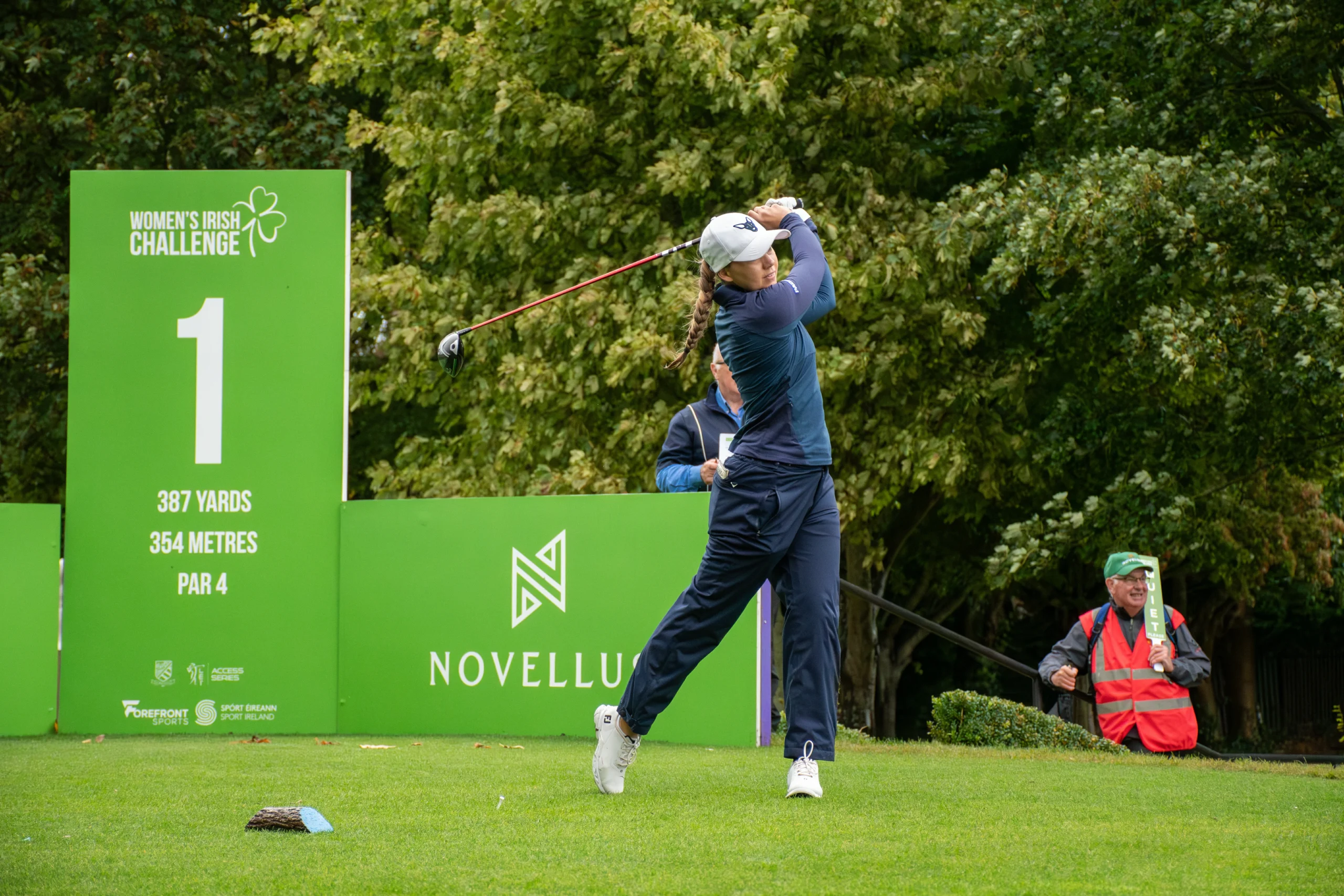 Female golfer in a navy outfit mid-swing on a green course, with a large green sign reading Women's Irish Challenge behind her.