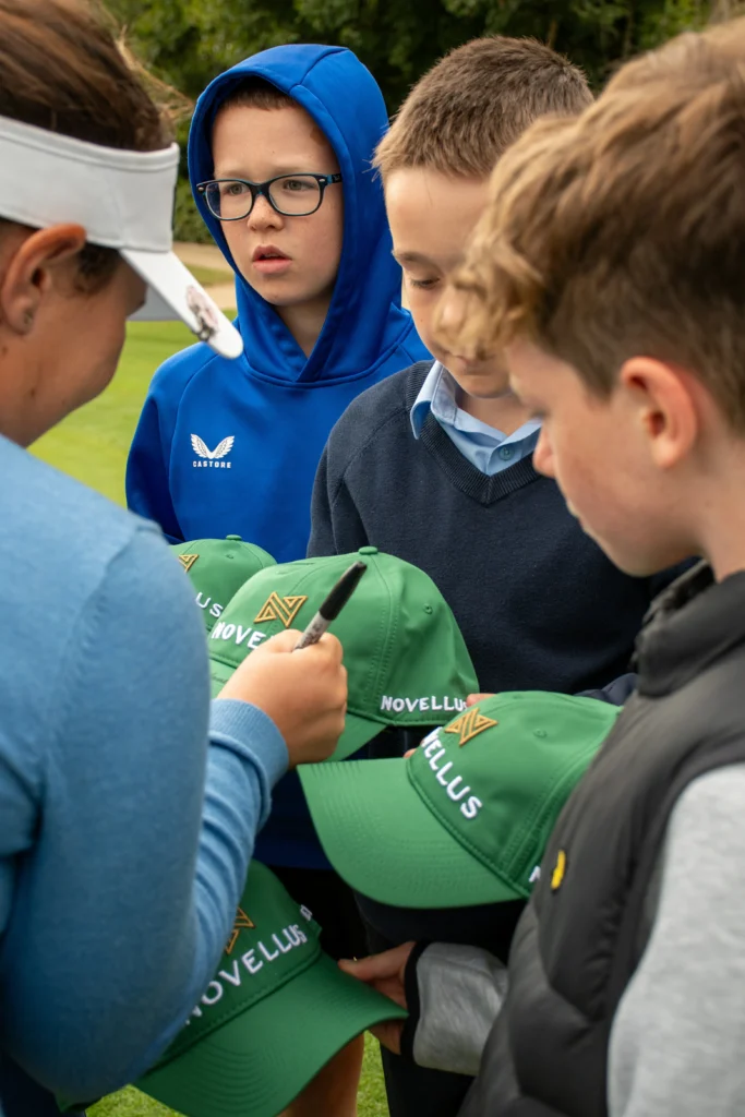 Group of youths gathered outdoors as one signs a green cap while others hold more caps. A boy in a blue hoodie with glasses watches nearby.