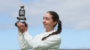 Woman with a braided ponytail raises a trophy outdoors, smiling at the camera against a blue sky.