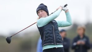 Female golfer in a dark beanie and navy vest mid-swing with a driver on a golf course.