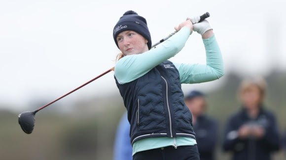 Female golfer in a dark beanie and navy vest mid-swing with a driver on a golf course.