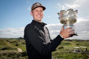 Man posing with a large silver trophy on a sunny golf course, wearing a dark jacket and cap.