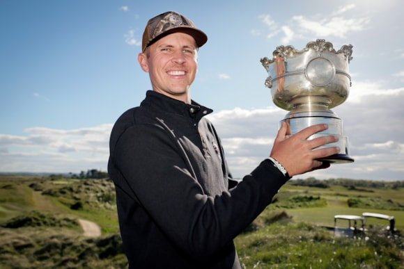 Man posing with a large silver trophy on a sunny golf course, wearing a dark jacket and cap.