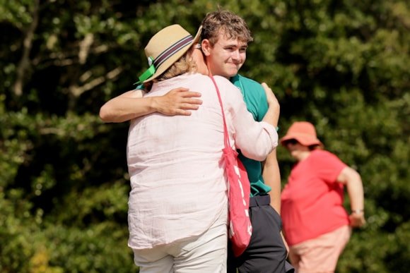 Young man in teal shirt hugs an older woman in light clothing outdoors; a person in red in the background observes.