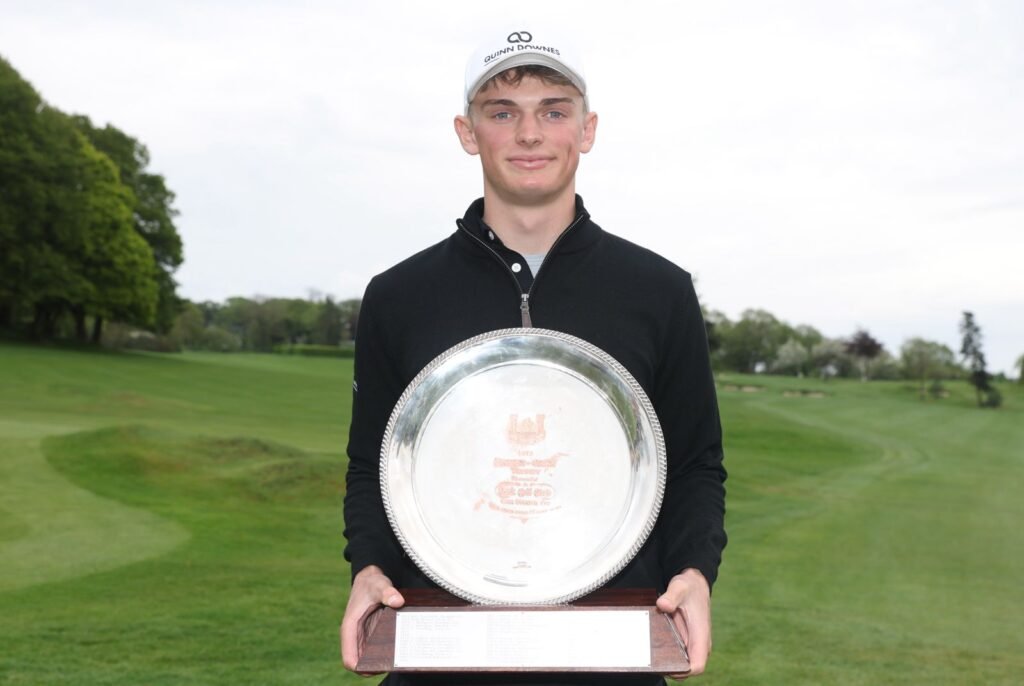 Young golfer on a green golf course holding a large silver trophy plate and a wooden base.