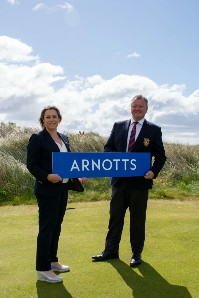 Two adults in business attire stand on a grassy area outdoors, holding a blue sign that reads ARNOTTS.