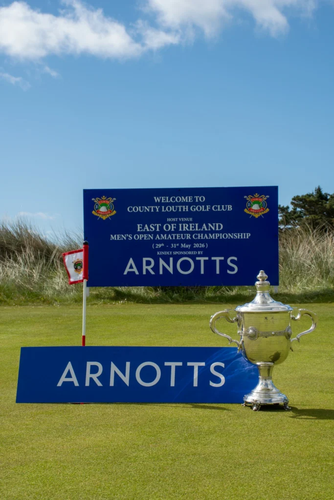 Blue event banner at a golf course for the East of Ireland Championship at County Louth Golf Club, with a large silver trophy in the foreground.