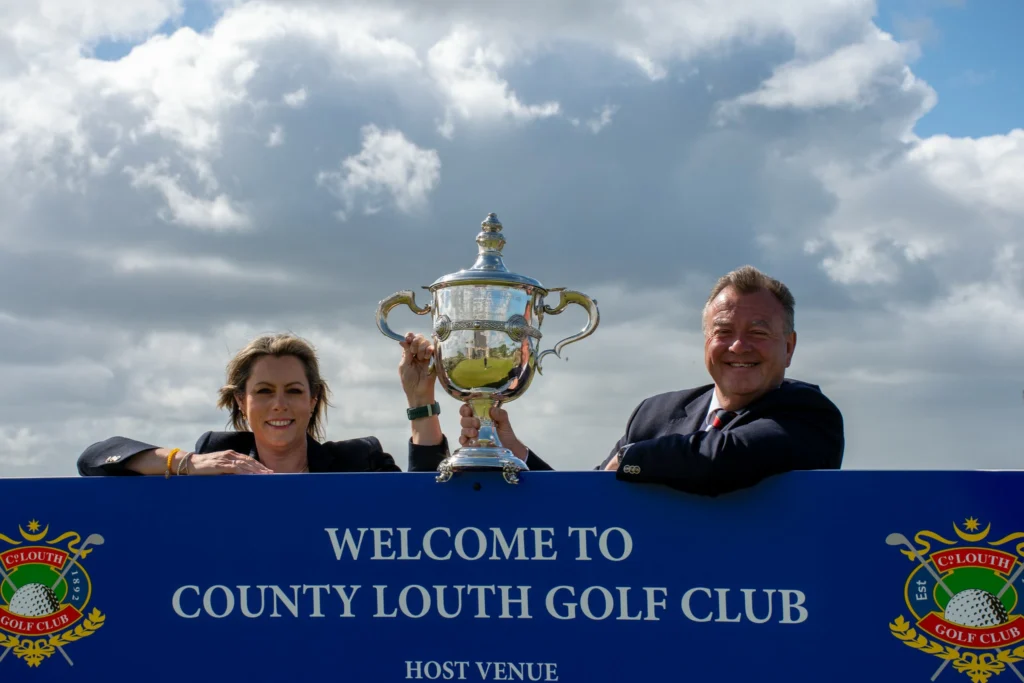 Two people in suits lift a large silver trophy, with a blue banner reading 'WELCOME TO COUNTY LOUTH GOLF CLUB' in the background.