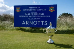Arnotts x East of Ireland-7 - Talk Golf Ireland - Talk Golf Ireland Blue sign for County Louth Golf Club event with a large silver trophy on a grassy course in front of it.