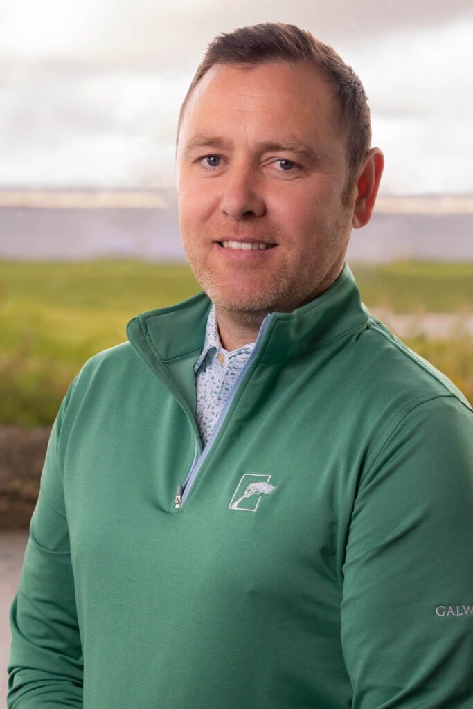 Brendan Mackey 2 - Talk Golf Ireland Portrait of a man outdoors wearing a green zip-up jacket, smiling at the camera with a blurred field in the background.