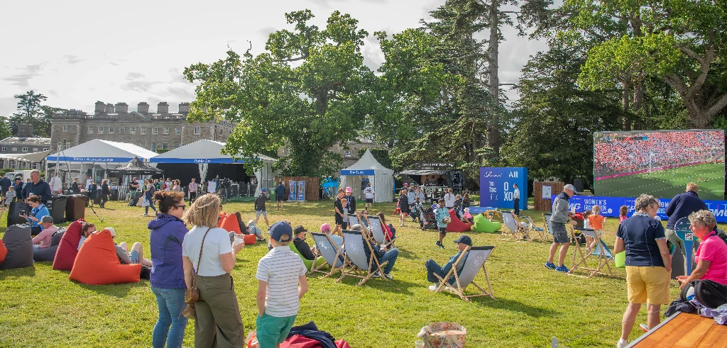 Outdoor park festival with tents, people lounging on bean bags and deck chairs, watching a large outdoor screen showing a sports match (crowd scene).