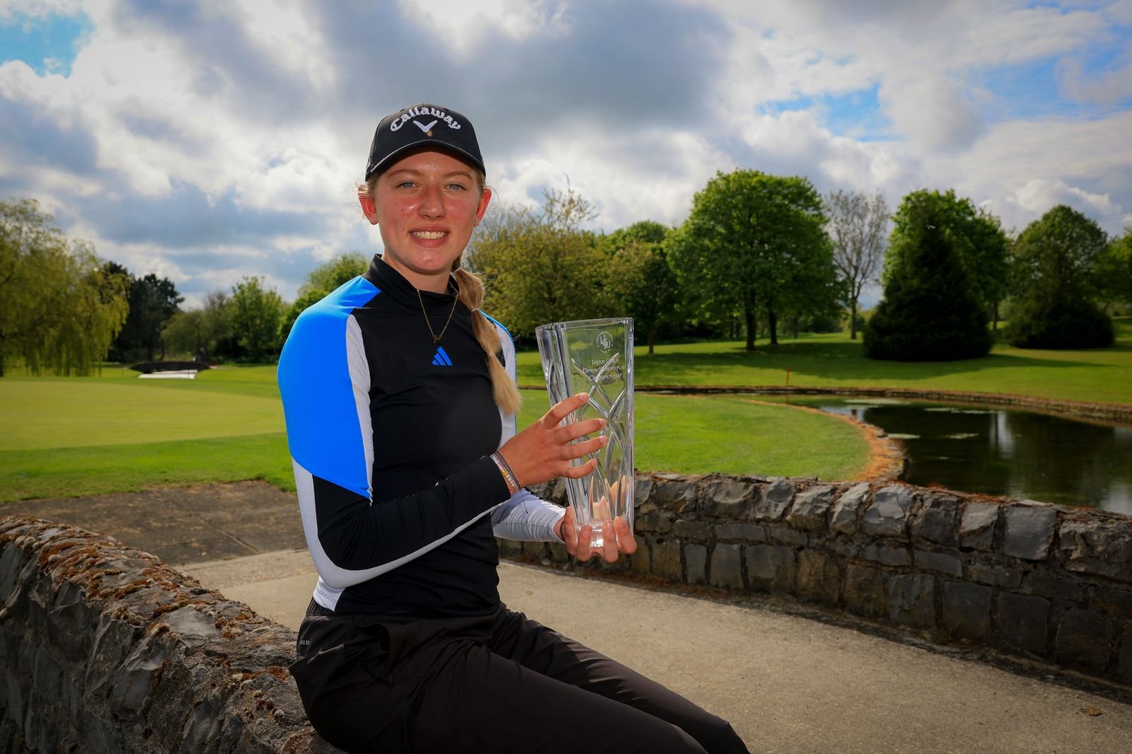 Young woman in a black and blue athletic top and Callaway cap sits on a stone wall, holding a tall crystal trophy outdoors on a golf course.