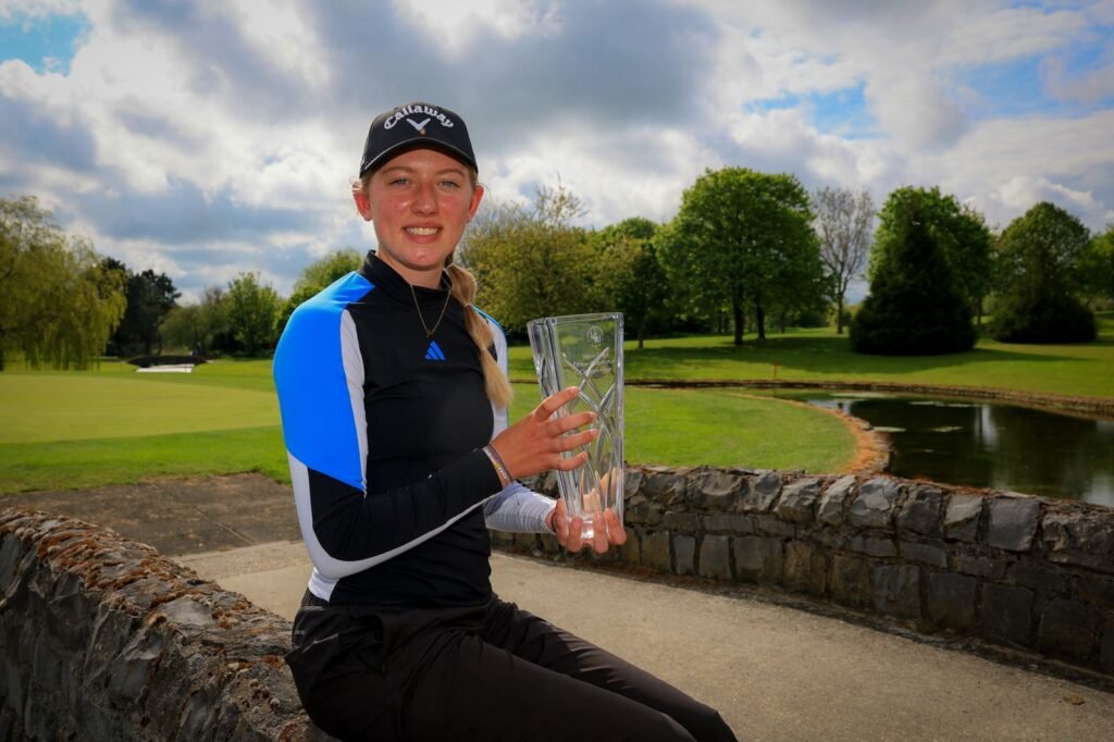 Young woman golfer wearing a Callaway cap, sitting on a stone railing and holding a tall glass trophy outdoors.