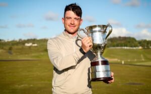 Man in light jacket holding a large silver trophy outdoors, smiling at the camera (sports victory)