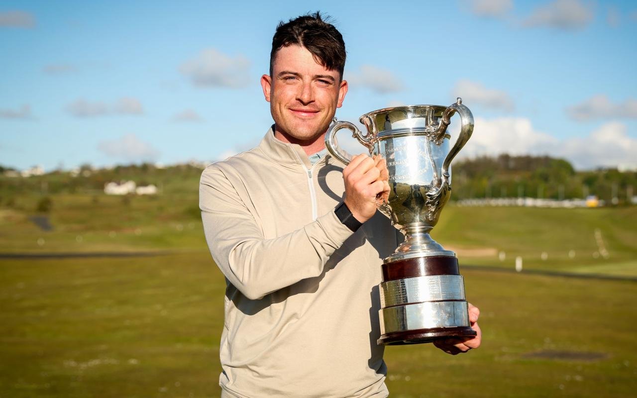 Man in light jacket holding a large silver trophy outdoors, smiling at the camera (sports victory)
