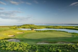 Coastal golf course with a green, sand bunkers, and water hazards under a blue sky.