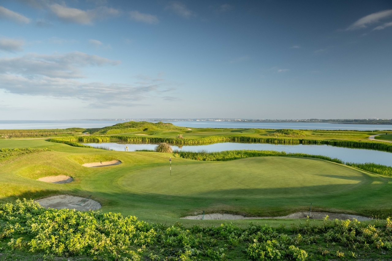 Coastal golf course with a green, sand bunkers, and water hazards under a blue sky.