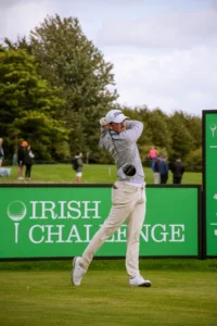 Male golfer in mid-swing on a golf course, with a bright green sign reading 'IRISH CHALLENGE' in the background.