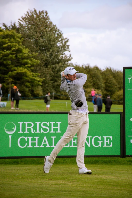 Male golfer in mid-swing on a golf course, with a bright green sign reading 'IRISH CHALLENGE' in the background.