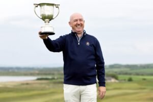 Smiling man in a navy jacket holds a large silver trophy outdoors against a cloudy sky and open landscape.