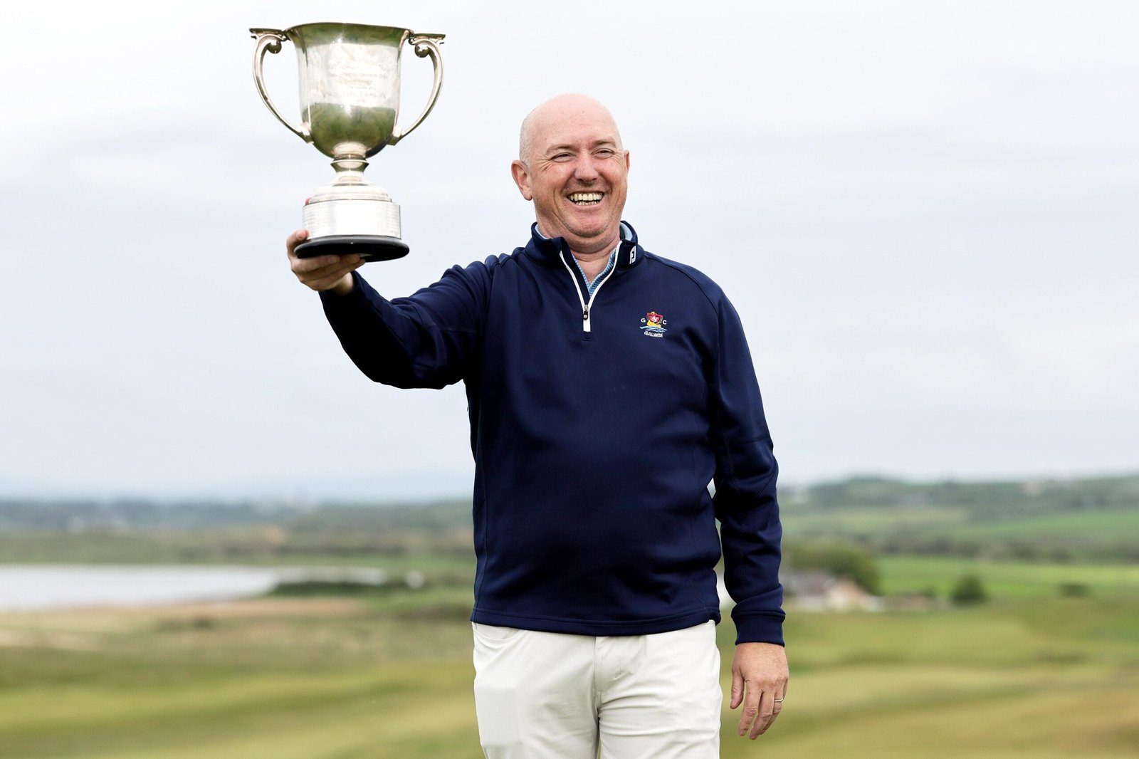 Smiling man in a navy jacket holds a large silver trophy outdoors against a cloudy sky and open landscape.