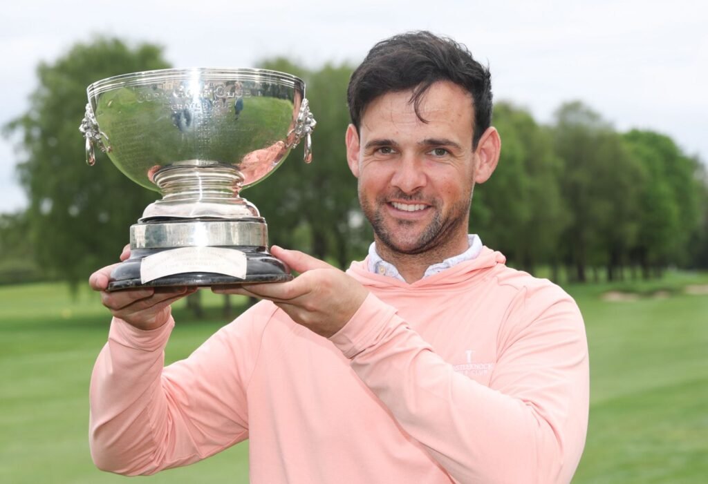 Man in a peach long-sleeve shirt holds a large silver trophy outdoors on a green golf course, smiling at the camera.