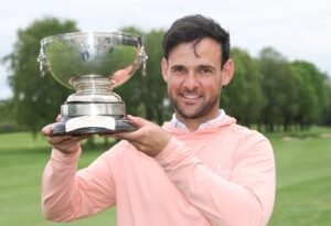 Man in a peach long-sleeve shirt holds a large silver trophy outdoors on a green golf course, smiling at the camera.