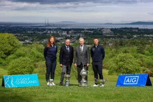 Four individuals pose with multiple trophies on a golf course, Golf Ireland and AIG banners in the foreground, with a coastal town and hills in the background.