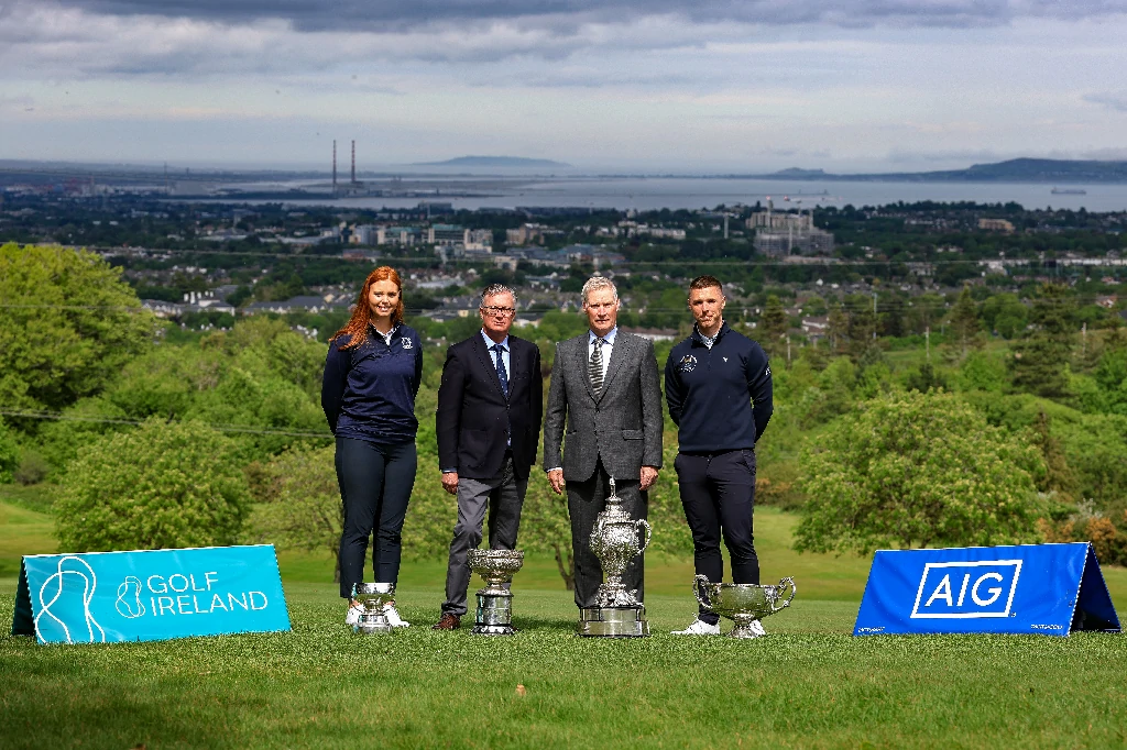 Four individuals pose with multiple trophies on a golf course, Golf Ireland and AIG banners in the foreground, with a coastal town and hills in the background.