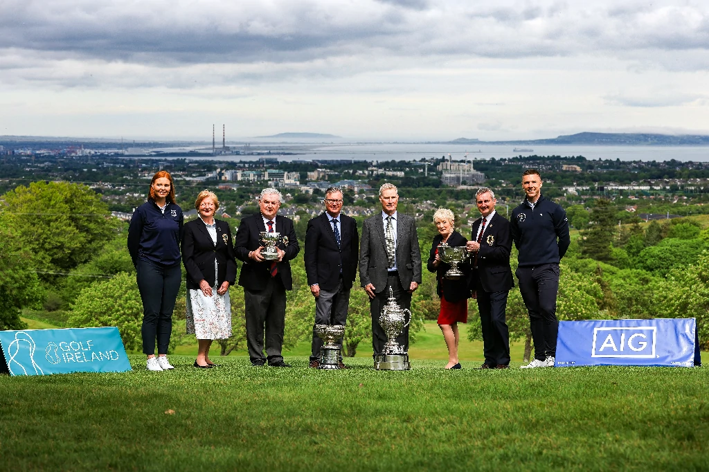 AIG launch 2 - Talk Golf Ireland Nine people on a golf course pose with three large silver trophies, banners in the foreground and a cityscape in the distance.