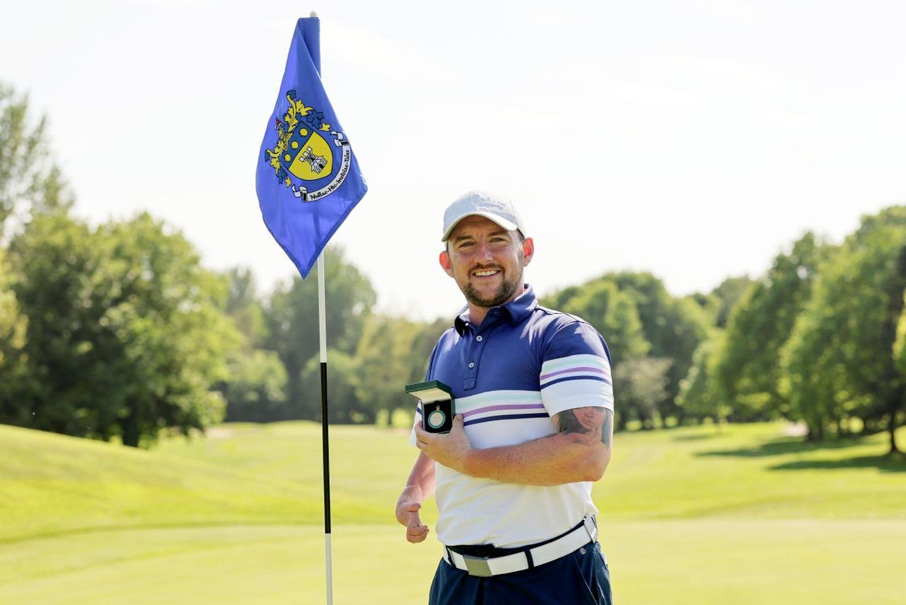 Smiling golfer in a blue striped polo holds a medal while standing near a blue flag on a sunny golf course crest in background.