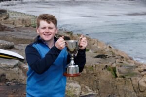 David Kitt 1 - Talk Golf Ireland Young man by the sea on a rocky shore, smiling while holding a silver trophy.