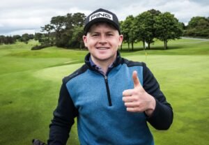 Man wearing a Ping cap smiles and gives a thumbs-up on a golf course.