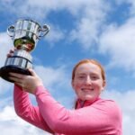 Girl in a pink long-sleeve top raising a large silver trophy against a blue sky, celebrating.