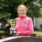 Young girl with red hair in a pink jacket holding a silver trophy outdoors, smiling at the camera.