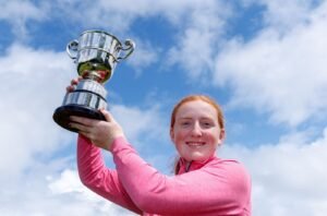 Girl in a pink long-sleeve top raising a large silver trophy against a blue sky, celebrating.