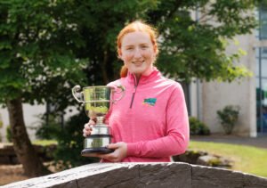 Young girl with red hair in a pink jacket holding a silver trophy outdoors, smiling at the camera.