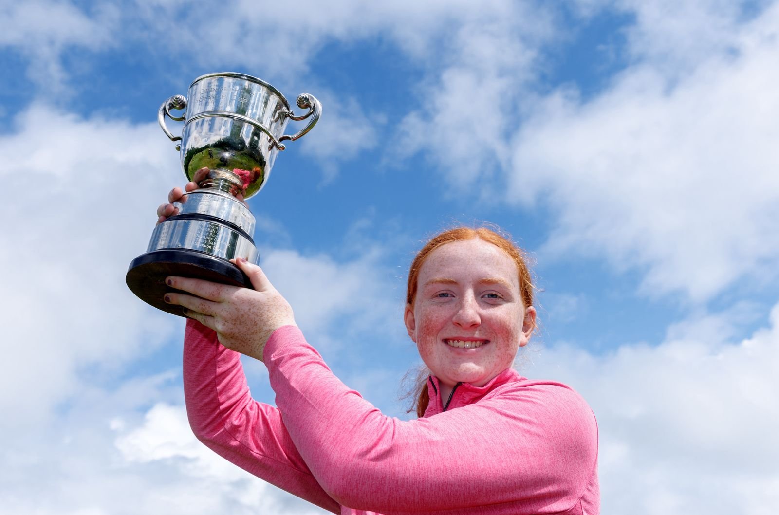 Girl in a pink long-sleeve top raising a large silver trophy against a blue sky, celebrating.