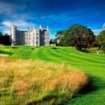 Castle-like stone building on a lush golf course under a bright blue sky with trees surrounding the grounds