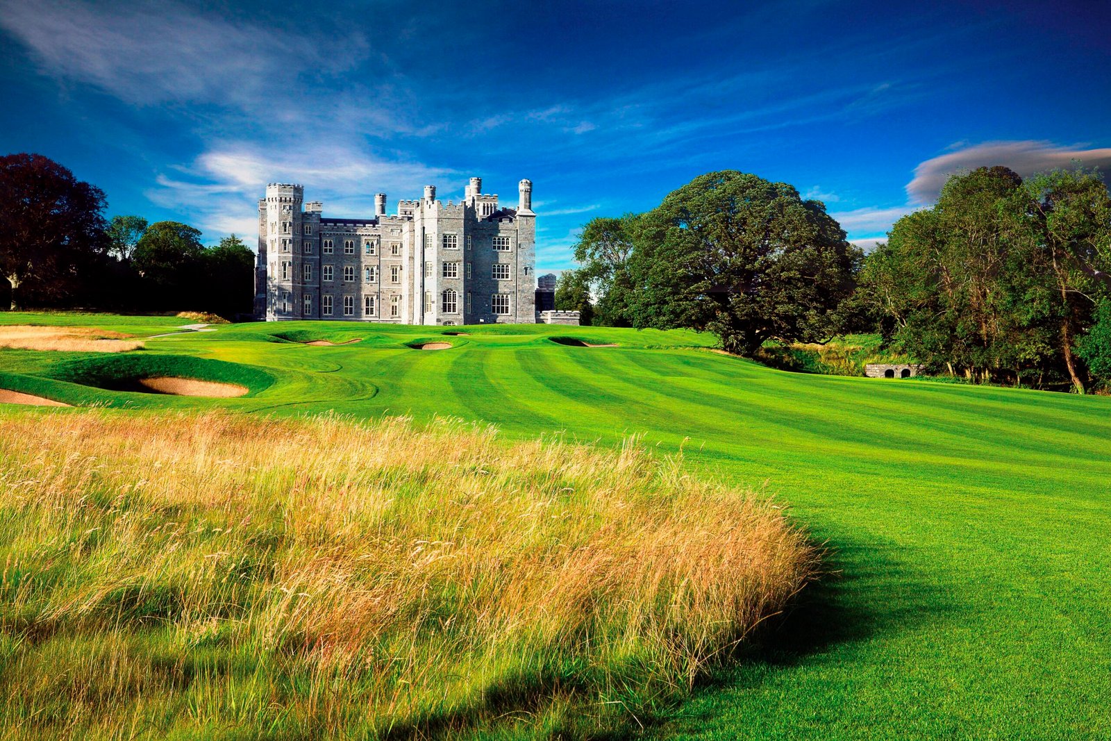 Castle-like stone building on a lush golf course under a bright blue sky with trees surrounding the grounds