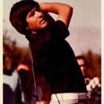 Man mid-swing playing golf, wearing a dark shirt and light plaid pants with spectators in the background.