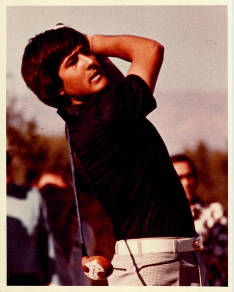 Man mid-swing playing golf, wearing a dark shirt and light plaid pants with spectators in the background.