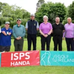 Eight people stand in a line on a golf course, posing for a group photo with colorful sponsor banners in front.