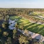 Aerial view of a golf course winding through a pine forest, with greens and sandy bunkers visible in the fairways and rough terrain nearby.
