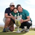 A smiling family of three holds a large silver golf trophy on a sunny course, posing for a celebratory photo.