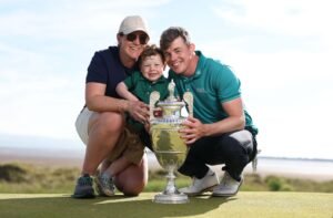 A smiling family of three holds a large silver golf trophy on a sunny course, posing for a celebratory photo.