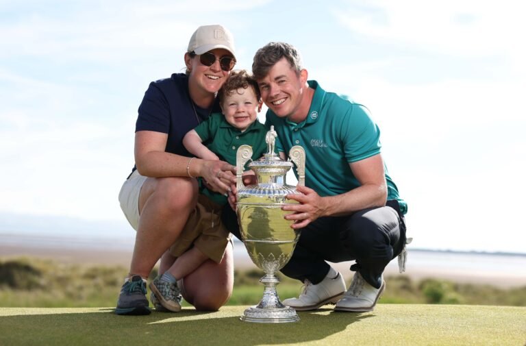 A smiling family of three holds a large silver golf trophy on a sunny course, posing for a celebratory photo.